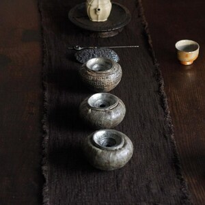 May include: An overhead shot of a tea ceremony setup. Three small, round, gray ceramic vessels with metal interiors are arranged on a dark brown runner. A small teacup, a teapot, and other tea ceremony tools are also visible.