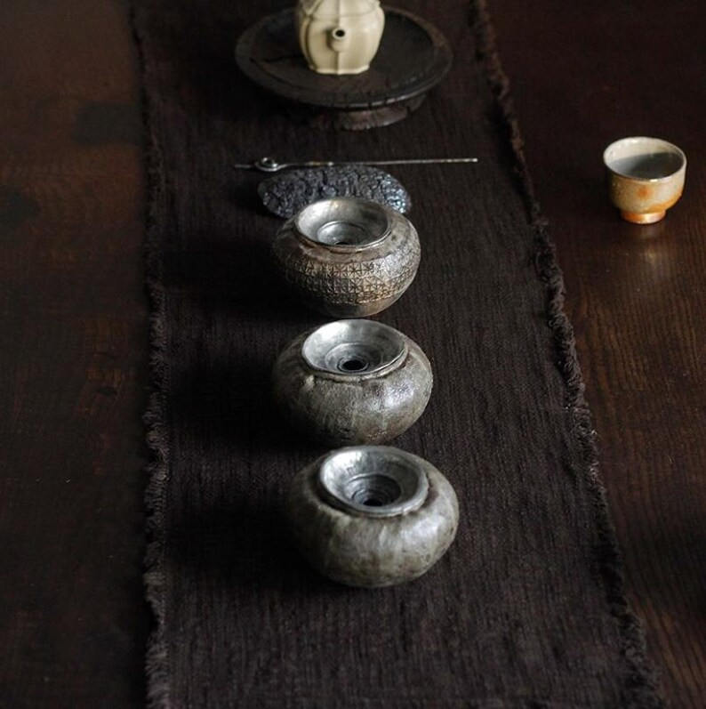 May include: An overhead shot of a tea ceremony setup. Three small, round, gray ceramic vessels with metal interiors are arranged on a dark brown runner. A small teacup, a teapot, and other tea ceremony tools are also visible.