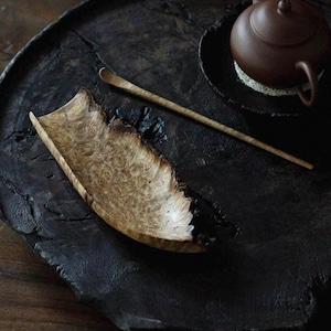 May include: A wooden tea set on a dark, textured surface. The set includes a light-colored, bark-edged wooden scoop, a long, thin wooden spoon, and a brown teapot. The items are arranged for a tea ceremony.