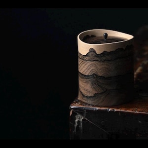 May include: A cylindrical wooden container with a lid. The container has a light beige rim and a dark brown, wood-grain pattern on the body. A small, dark knob sits atop the lid. The container is on a dark wooden surface.
