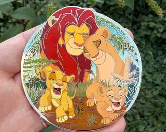 May include: A circular enamel pin featuring a colorful illustration of a lion family. The design depicts two adult lions and two playful lion cubs in a savanna setting with green foliage, a blue sky, and brown earth, rendered in a vibrant cartoon style.