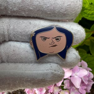 May include: A pin featuring a cartoon character with blue hair, a light skin tone, and a stern expression. The pin has a gold outline and is set against a gray background with pink flowers and green foliage.
