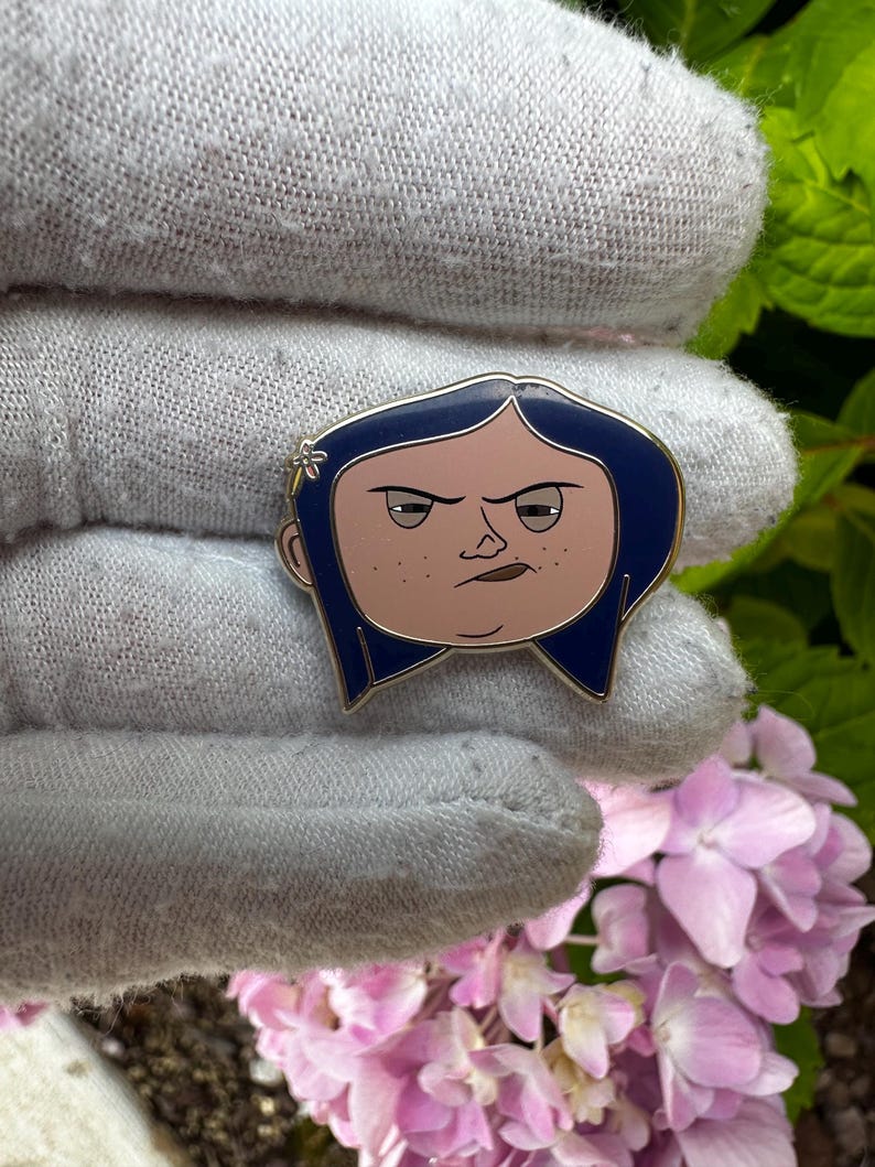 May include: A pin featuring a cartoon character with blue hair, a light skin tone, and a stern expression. The pin has a gold outline and is set against a gray background with pink flowers and green foliage.