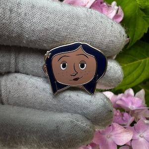 May include: Enamel pin featuring a cartoon character with dark blue hair, brown skin, and a small flower in her hair. The pin has a gold outline and is set against a background of pink flowers and green leaves.