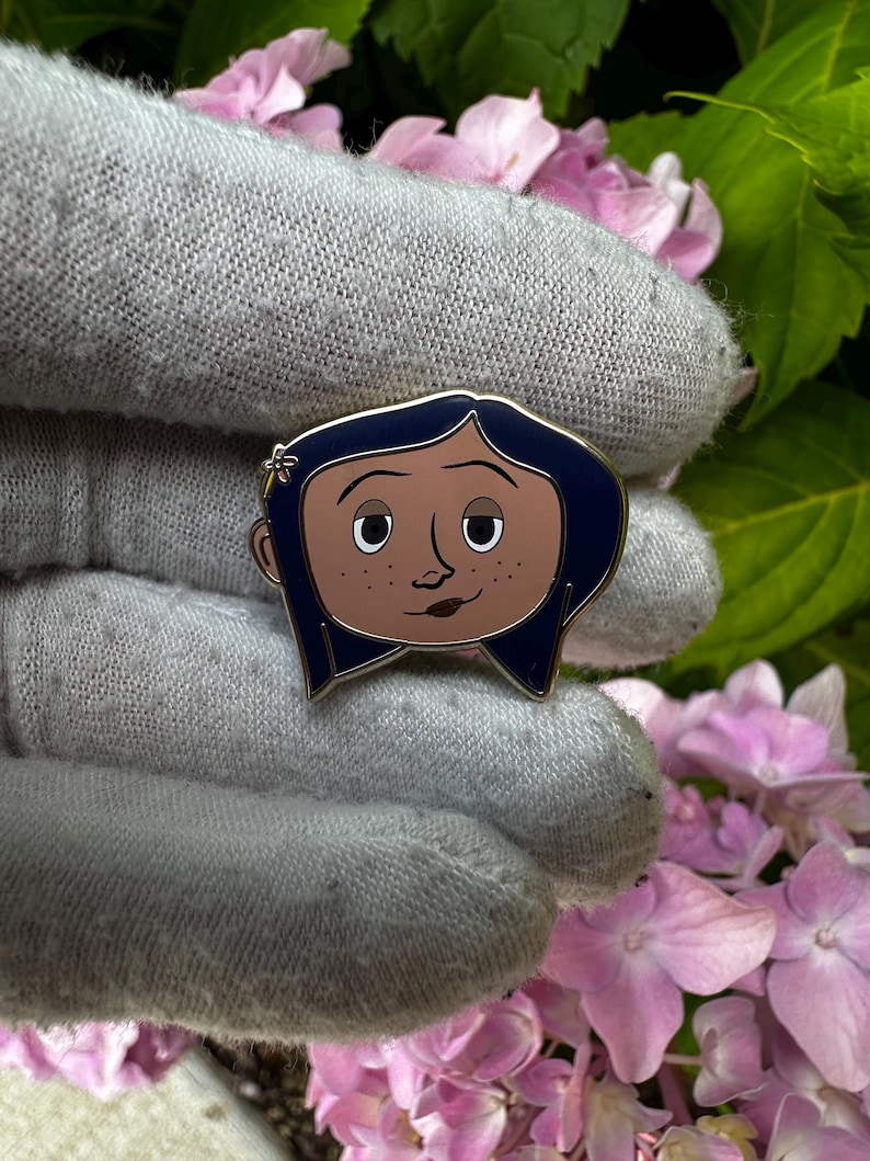 May include: Enamel pin featuring a cartoon character with dark blue hair, brown skin, and a small flower in her hair. The pin has a gold outline and is set against a background of pink flowers and green leaves.