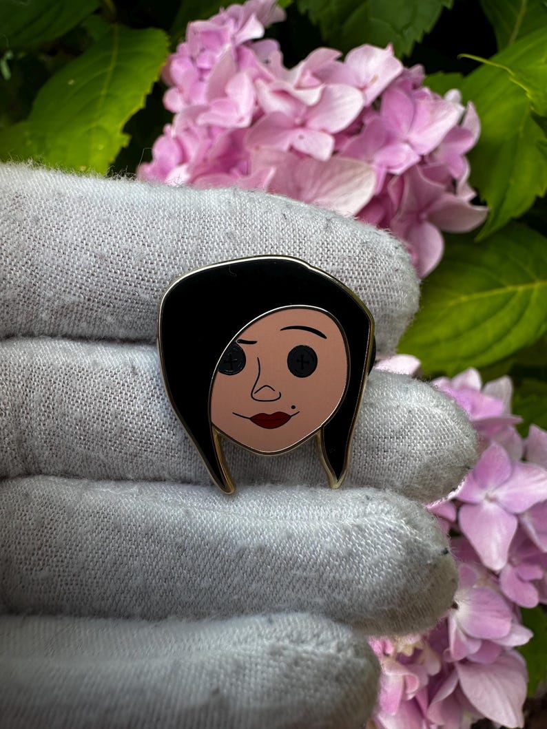 May include: A black enamel pin featuring a cartoon character with black hair, button eyes, and red lips. The pin has a gold border and is held against a backdrop of pink flowers and a gray glove.