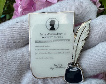 May include: A gold-toned pin featuring a parchment with the text "Lady Whistledown's SOCIETY PAPERS" and a silhouette. A white feather quill and black inkwell are also present. The pin is held against a backdrop of pink flowers.