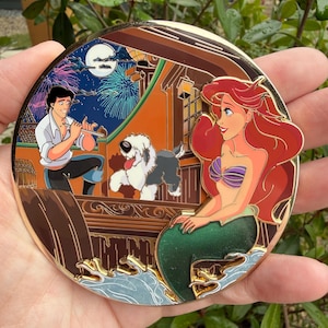 May include: A circular pin with a gold border, depicting a scene from The Little Mermaid. Ariel is on the right, Prince Eric plays a flute on the left, and a dog is in the middle, with fireworks in the background.