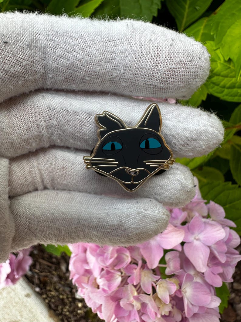 K&ouml;nnte beinhalten: Ein schwarzer Katzen-Emaille-Pin mit goldfarbenem Rand und leuchtend blauen Augen. Der Pin zeigt ein stilisiertes Katzengesicht-Design. Der Pin wird in einem grauen Handschuh gehalten, mit rosa Hortensienbl&uuml;ten und gr&uuml;nen Bl&auml;ttern im Hintergrund.