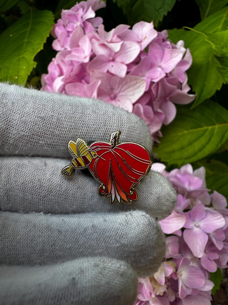 May include: A red and gold enamel pin shaped like a stylized apple with a yellow and gold bee. The pin is set against a gray background with pink flowers and green leaves in the background.