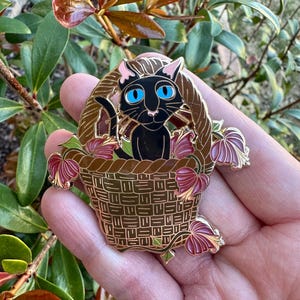 May include: A gold-colored enamel pin featuring a black cat with blue eyes sitting in a woven basket. The basket is surrounded by burgundy and gold-colored leaves. The pin is held in a hand against a background of green and brown foliage.