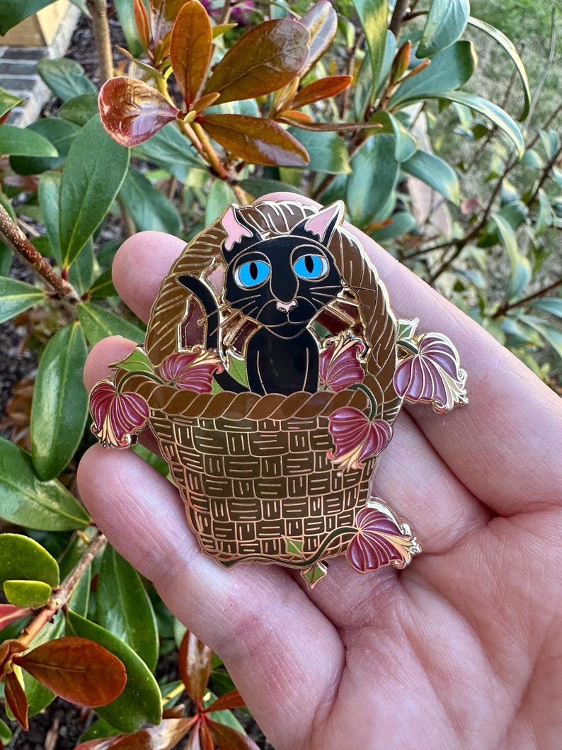May include: A gold-colored enamel pin featuring a black cat with blue eyes sitting in a woven basket. The basket is surrounded by burgundy and gold-colored leaves. The pin is held in a hand against a background of green and brown foliage.