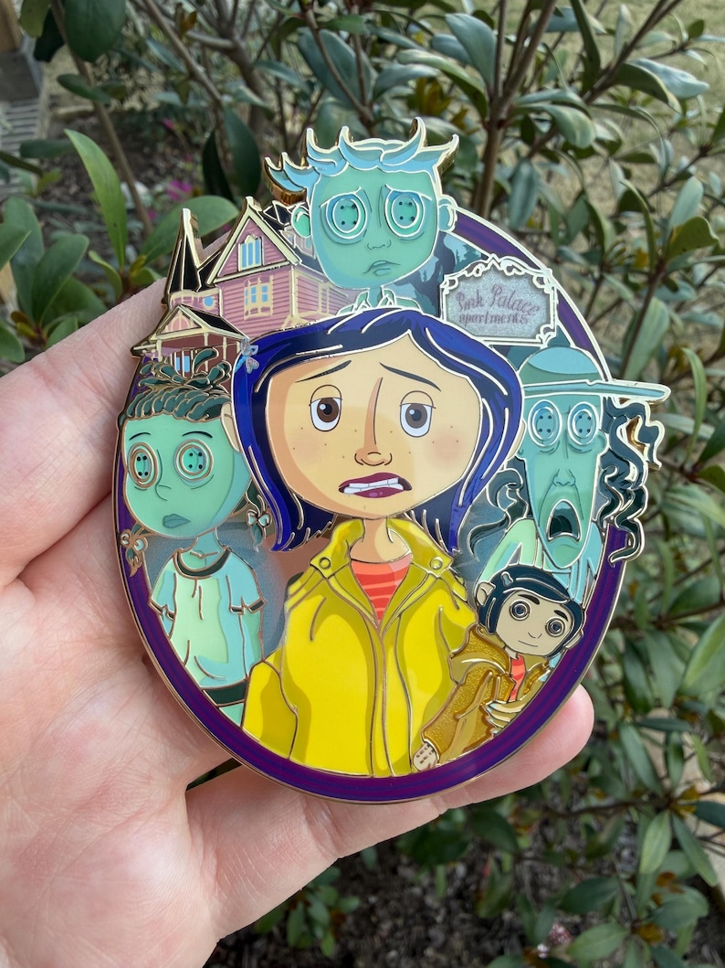 May include: A round, purple enamel pin featuring characters from the animated film, Coraline. The pin includes images of Coraline, the other mother, and other characters. The words "Pink Palace Apartments" are also visible on the pin.