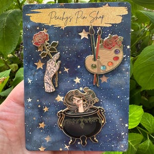 Puede incluir: Colección de pines esmaltados sobre un fondo azul marino con estrellas doradas. Los pines incluyen una rosa sostenida por una mano, una paleta de pintor con pinceles y una bruja en un caldero. El texto "Peachys Pin Shop" está en la parte superior.