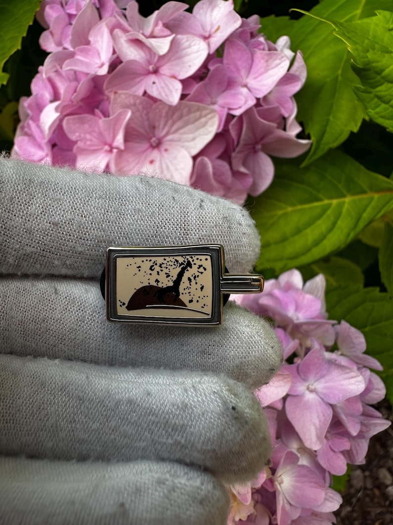 K&ouml;nnte beinhalten: Rechteckiger silberfarbener Manschettenknopf mit einem schwarz-braunen Grafikdesign. Das Design scheint eine Silhouette eines Tieres zu sein. Der Manschettenknopf wird vor einem Hintergrund aus rosa Bl&uuml;ten und gr&uuml;nen Bl&auml;ttern gehalten.