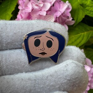 May include: Enamel pin featuring a cartoon character with blue hair, black button eyes, and a light pink face with freckles. The pin has a silver outline and a small flower detail. The background includes pink flowers and green leaves.