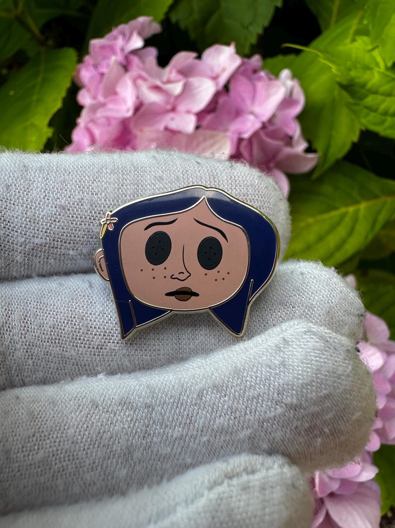 May include: Enamel pin featuring a cartoon character with blue hair, black button eyes, and a light pink face with freckles. The pin has a silver outline and a small flower detail. The background includes pink flowers and green leaves.