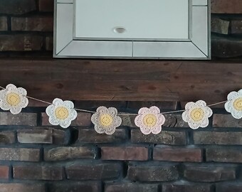 Op de afbeelding: Een slinger van gehaakte bloemen in wit, roze en geel, geregen aan een dun koord. De bloemen zijn in een rij gerangschikt, waardoor een decoratieve slinger ontstaat. De slinger wordt getoond tegen een donker houten oppervlak en een bakstenen muur.
