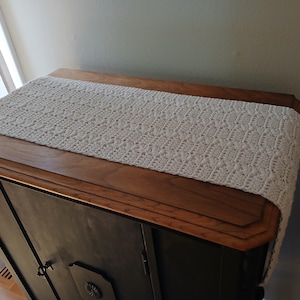 Op de afbeelding: Een crèmekleurige, gehaakte tafelloper met een gedetailleerd open patroon. De loper is rechthoekig en ligt op een donkere houten kast met een lichtere houten rand. De loper voegt een vleugje handgemaakte charme toe.