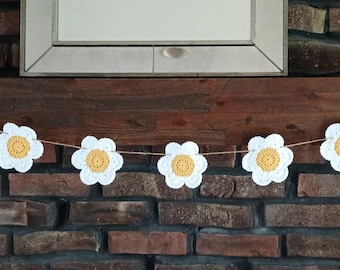 Op de afbeelding: Een handgemaakte gehaakte madeliefjesslinger met witte bloemblaadjes en gele centra. De slinger is geregen over een donkere houten mantel voor een bakstenen muur. Decoratieve woondecoratie.