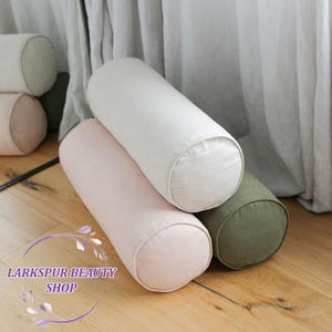 Puede incluir: Se muestran varios cojines cilíndricos en varios colores. Los cojines son en tonos blanco, rosa y verde. Están hechos de una tela suave y tienen extremos redondeados. La imagen también incluye el texto "Larkspur Beauty Shop".