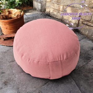 Peut inclure: Un coussin de méditation rond rose poussiéreux avec un tissu texturé. Le coussin est posé sur une surface en pierre grise. Le texte "Larkspur Beauty Shop" est dans le coin supérieur droit.