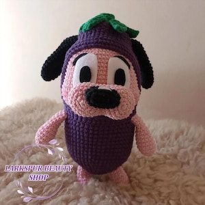 Puede incluir: Un peluche de crochet hecho a mano con forma de perro con un disfraz de berenjena. El juguete es principalmente morado, rosa, negro y verde. El texto "Larkspur Beauty Shop" es visible en la parte inferior izquierda.