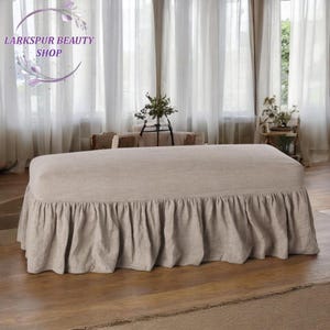 May include: A beige upholstered ottoman with a ruffled skirt. The rectangular ottoman sits on a wooden floor. The background features sheer white curtains and decorative plants. The shop logo "Larkspur Beauty Shop" is in the upper left corner.