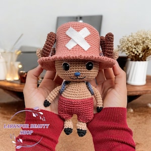 May include: A handmade crochet doll of a cartoon character, featuring a pink hat with a white cross, brown antlers, and red shorts. The doll is held in front of a blurred background. The doll has a blue nose and black eyes. The text "Larkspur Beauty Shop" is visible.