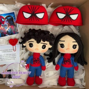 May include: A handmade crochet set featuring two Spider-Man themed dolls, two red hats with white eye designs, and a heart-shaped paper clip. The dolls are dressed in blue and red outfits. The text "LARKSPUR BEAUTY SHOP" is visible.