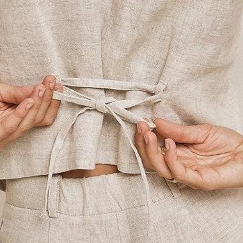 May include: Close-up of a beige linen top with a tie-back detail. The top is being tied into a bow by a person's hands. The fabric appears to be lightweight and textured, with a natural, neutral colour. The bottom of the top is visible, along with the top of the matching trousers.
