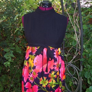 May include: A black sleeveless dress with a high neckline and a floral print skirt. The skirt features large pink, yellow, and green flowers on a dark blue background. A black tie belt cinches the waist.