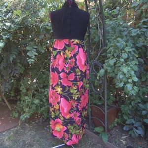 May include: A vintage maxi dress on a mannequin. The dress has a black bodice and a vibrant floral print skirt with pink, yellow, and green flowers on a black background. The dress is displayed outdoors with greenery in the background.