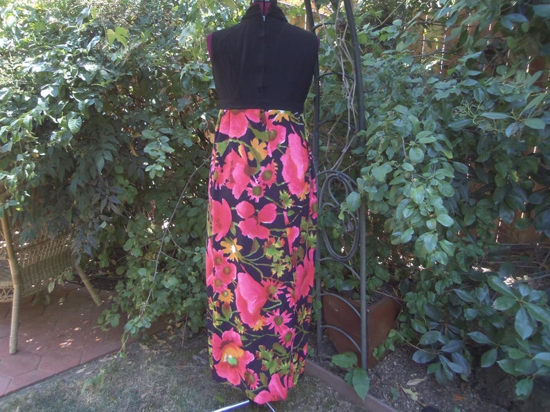 May include: A vintage maxi dress on a mannequin. The dress has a black bodice and a vibrant floral print skirt with pink, yellow, and green flowers on a black background. The dress is displayed outdoors with greenery in the background.