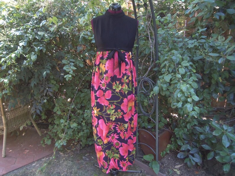 May include: A vintage maxi dress with a black bodice and a vibrant floral skirt. The skirt features large pink and yellow flowers on a dark background. A black tie belt cinches the waist. The dress is displayed on a mannequin.