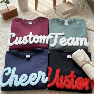 Può includere: Quattro felpe girocollo bordeaux, verde salvia, blu navy e nere. Ogni felpa ha una parola soffice e strutturata sul davanti: "Custom", "Team", "Cheer" e "Custom". Sono visibili anche un asciugamano arrotolato, occhiali e una piccola pianta.