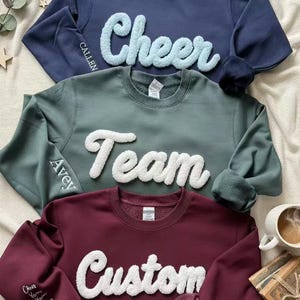 May include: Three crewneck sweatshirts in navy, sage green, and burgundy. Each features a fluffy, white word: "Cheer", "Team", and "Custom". The navy sweatshirt has "Callen" embroidered on the sleeve. The burgundy sweatshirt has a heart.