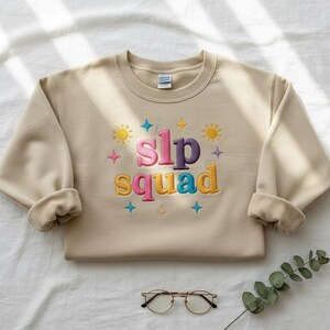 May include: A beige crewneck sweatshirt with the embroidered text "slp squad" in colorful letters. The design includes sun and star accents. The sweatshirt is folded, with a pair of glasses and a sprig of greenery in the foreground.