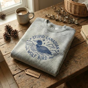 Puede incluir: Una sudadera gris con un gráfico azul y blanco de un ave colimbo y el texto "STUPID CANADIAN WOLF BIRD". La sudadera está doblada sobre una mesa de madera junto a una taza de café, piñas y una pila de libros.