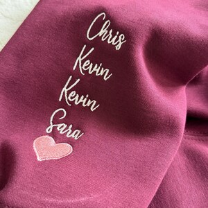 May include: Burgundy-colored sweatshirt sleeve with white embroidered names: Chris, Kevin, Kevin, and Sara. A pink heart is embroidered below Sara's name. The fabric is a solid color and appears to be a soft material.