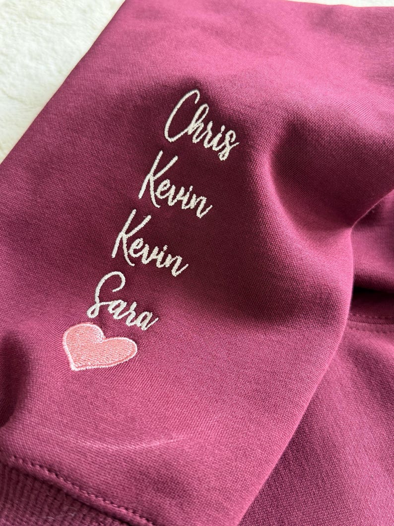 May include: Burgundy-colored sweatshirt sleeve with white embroidered names: Chris, Kevin, Kevin, and Sara. A pink heart is embroidered below Sara's name. The fabric is a solid color and appears to be a soft material.