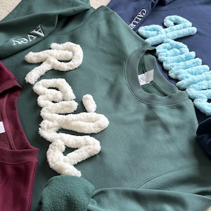 May include: A collection of crewneck sweatshirts in various colors, including teal, navy, and burgundy. Each sweatshirt features a name embroidered or appliqu&eacute;d in a contrasting color. The names are in a cursive font, and the sweatshirts are made of a soft material.