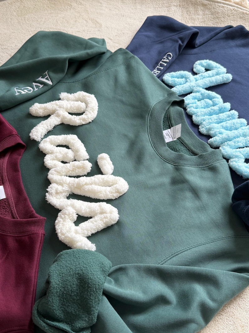 May include: A collection of crewneck sweatshirts in various colors, including teal, navy, and burgundy. Each sweatshirt features a name embroidered or appliqu&eacute;d in a contrasting color. The names are in a cursive font, and the sweatshirts are made of a soft material.