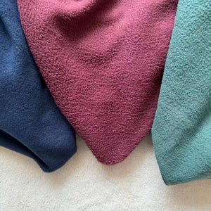 May include: Close-up of three pieces of textured fabric in shades of navy blue, burgundy, and teal. The fabrics are arranged on a white, textured surface, with the colors overlapping slightly. The texture appears soft and plush.