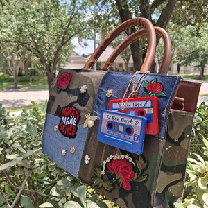 Peut inclure: Un sac à main patchwork composé de panneaux en camouflage et en jean avec des anses marron. Il est orné d'écussons de roses rouges, de petits clous floraux, d'une broche abeille et d'un écusson MAKE IT MAKE SENSE. Deux cassettes pendent.