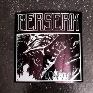 May include: Square sticker with the word "BERSERK" in bold, outlined white letters at the top. The lower portion features a black and white illustration of a character's face with sharp features and a dark background.