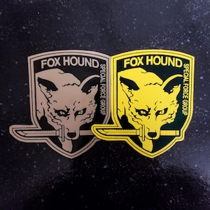 Peut inclure: Deux logos Foxhound Special Force Group. L'un est noir, jaune et vert. L'autre est noir, beige et vert.