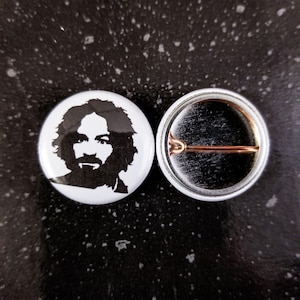 May include: A white button with a black and white silhouette of a man's face. The back of the button has a metal pin and a safety clasp.