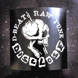 Könnte beinhalten: Schwarz-weißer Aufkleber mit einem Totenkopf- und Knochen-Design. Der Text um den Schädel lautet "D-Beat Rad Punk Disclose".