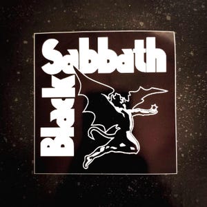 以下が含まれることがあります： 黒い背景に、翼と尻尾のある白い悪魔の図柄が描かれたブラック・サバスのロゴステッカーです。